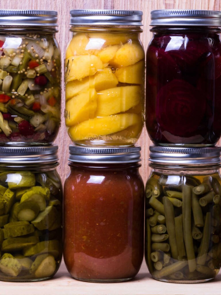 6 canning jars top row filled with beans, peaches, beets, the bottom row has pickles, tomato sauce, green beans.