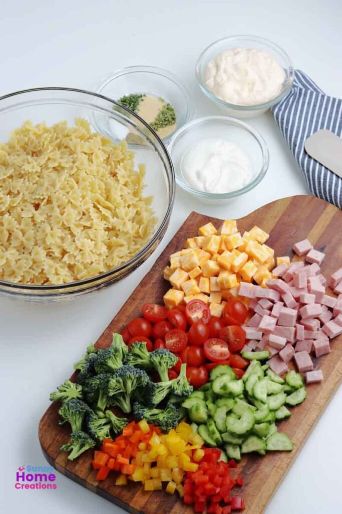 bowl of bowtie pasta noodles, small bowl of seasoning, small bowl of sour cream, small bowl of mayonnaise, cutting board with cut up cheese, ham, cherry tomatoes, cucumbers, bell peppers, and broccoli on it.