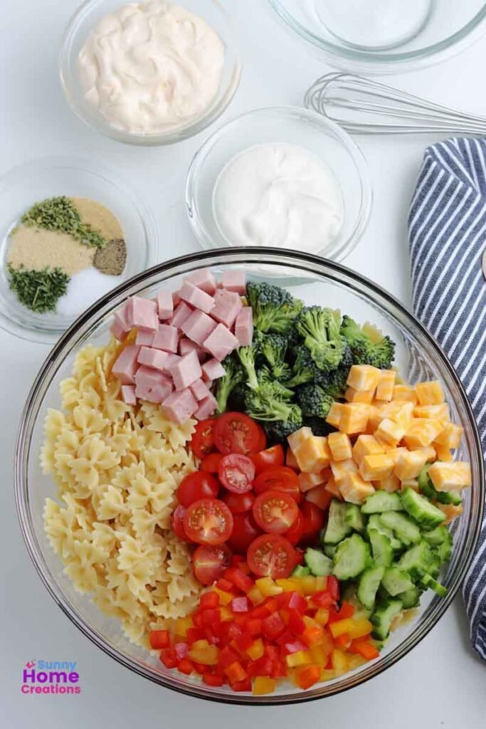 small bowl of mayonnaise, small bowl of sour cream, small bowl of spices, and bowl with diced ham, broccoli, cheese, cucumbers, bell peppers, bow tie noodles, and cherry tomatoes in it.