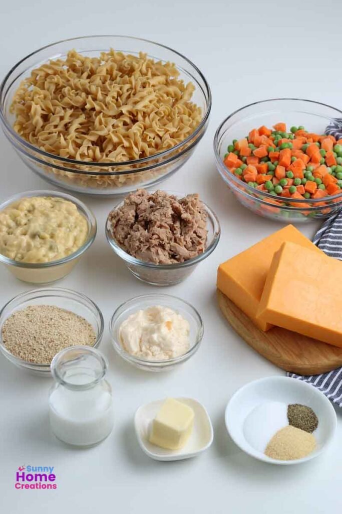 top left and around to the right and down - bowl of egg noodles, bowl of peas & carrots, two chunks of cheese on a small cutting board, small bowl with seasonings, small bowl with butter, small container of milk, bowl of bread crumbs, bowl of cream of celery, bowl of tuna, bowl of mayonaise.