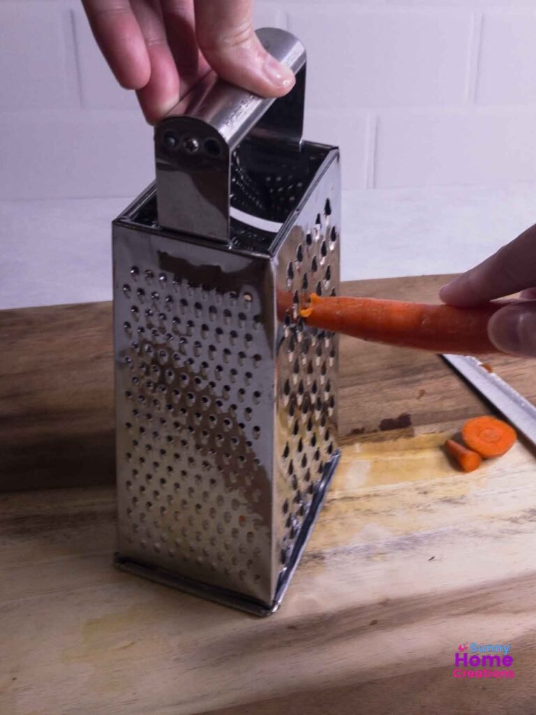 hand holding box grater handle with other hand grating carrot.
